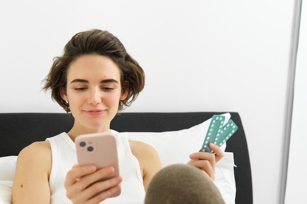 Free photo portrait of woman in bed holding mobile phone and pills taking medication searching for dosage and